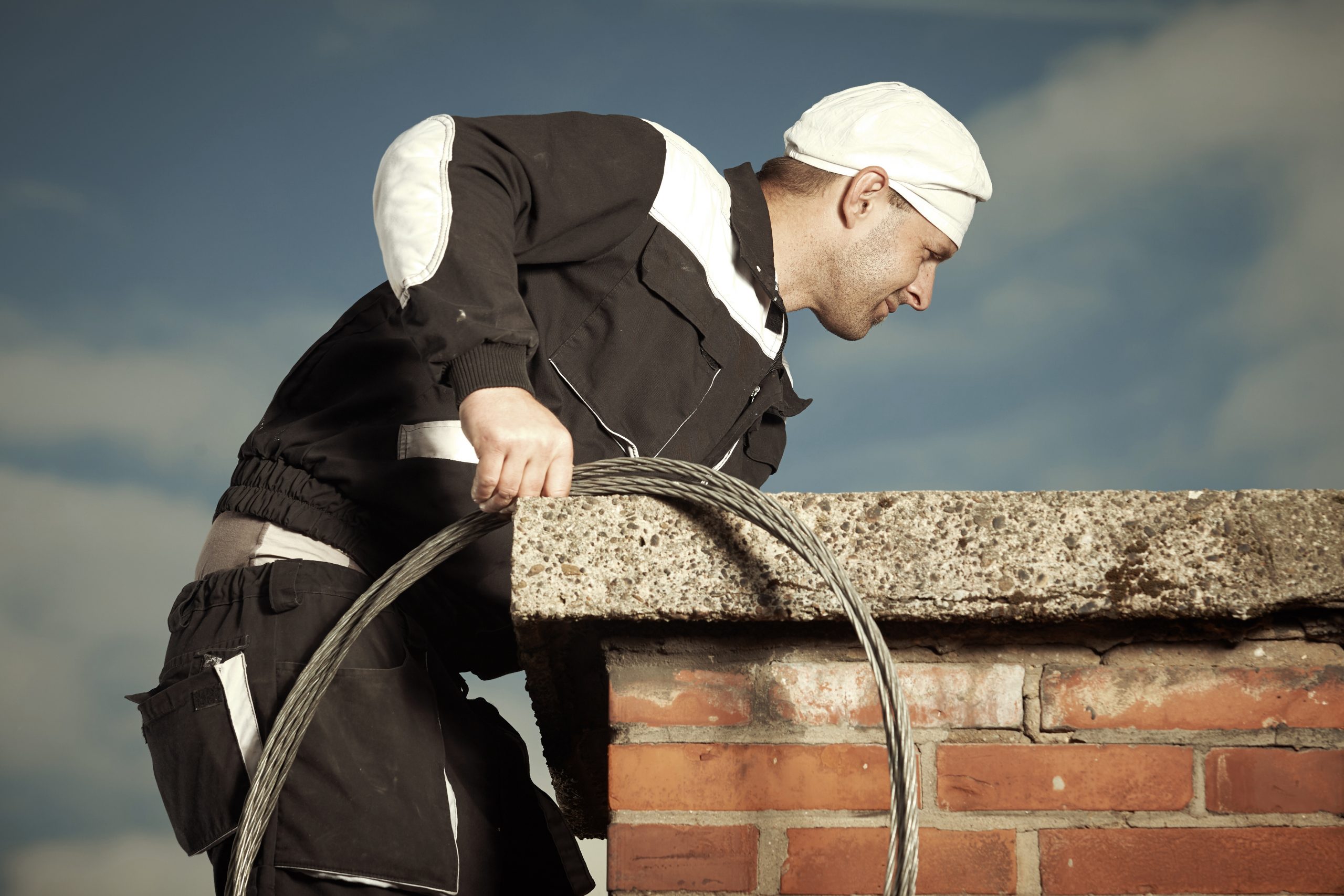 Chimney sweep man in work uniform cleaning brick style chimney on building roof